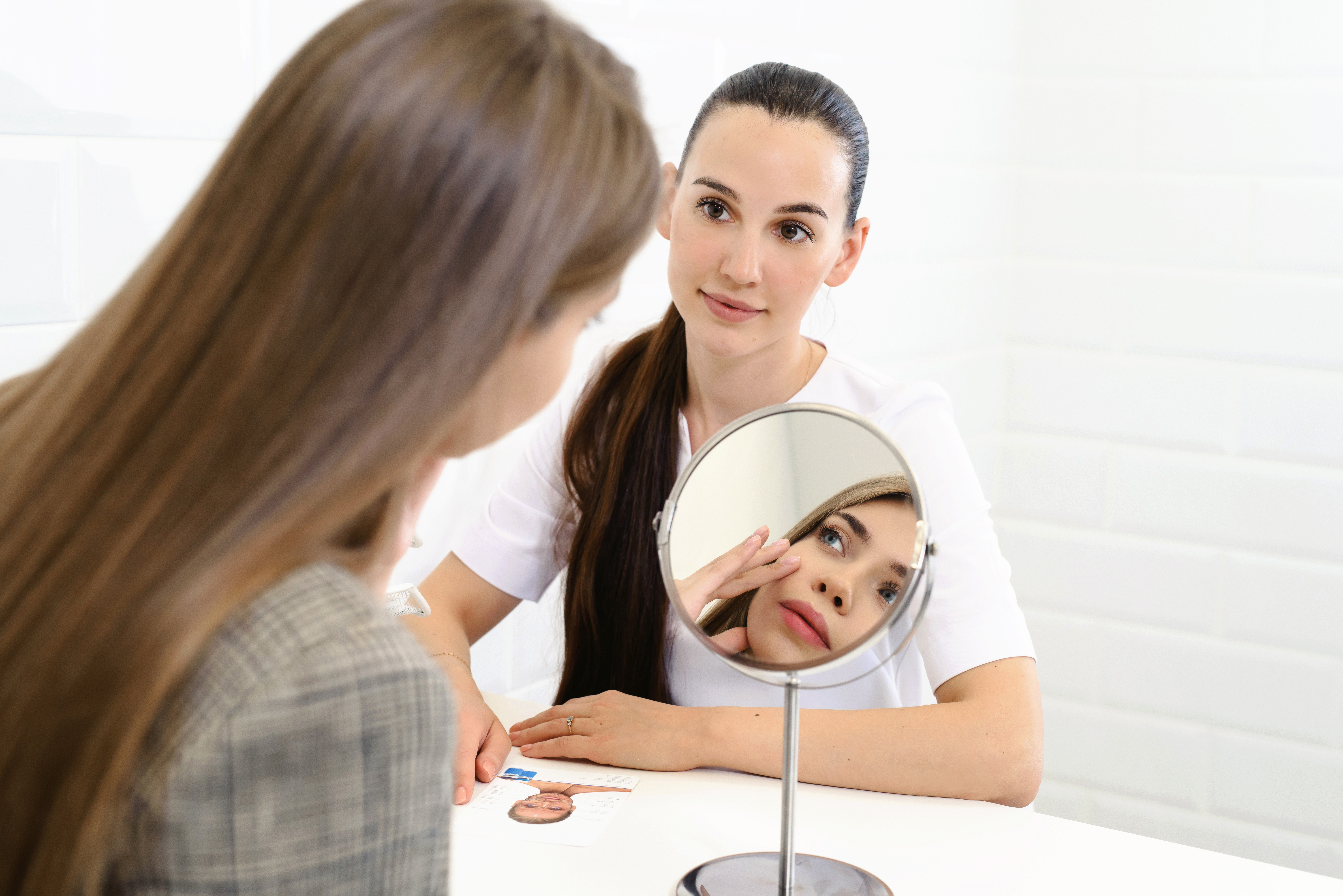 Consultation in cosmetology clinic. Female beauty doctor talking with patient. A young girl tells what she wants change in her face. Client shows on himself using mirror. Doctor listening carefully